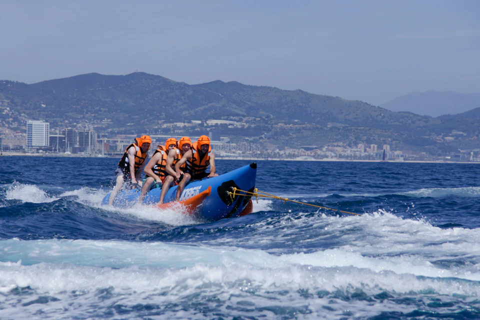 Four people wearing orange helmets and life vests are riding a blue inflatable banana boat on the ocean, with mountains in the background.