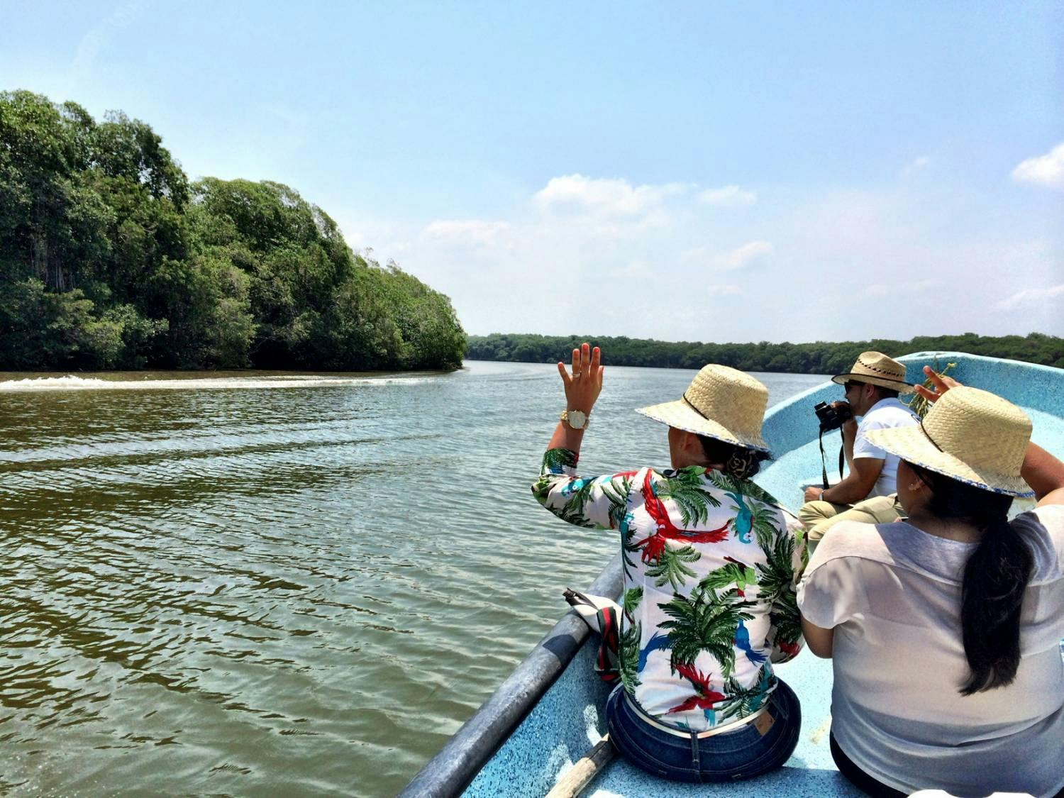 People in hats on a boat ride through a river, waving at the green, forested shoreline under a clear, sunny sky.