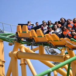 Människor som åker berg- och dalbana med gröna spår och gula stöd, skriker och ler mot en klarblå himmel.