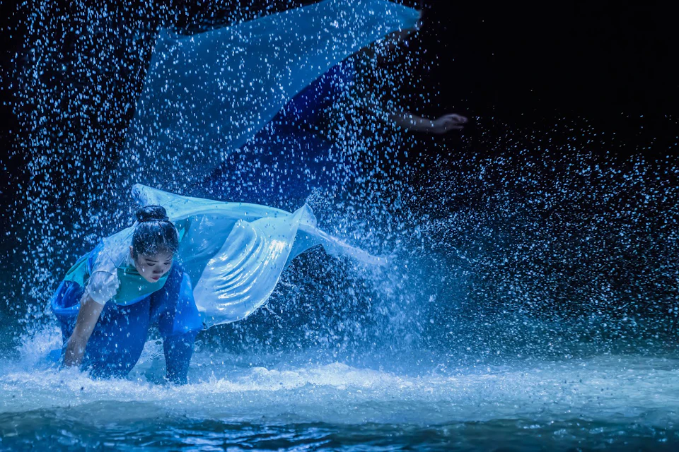 A performer in a flowing blue costume dances amid splashing water under dramatic lighting.