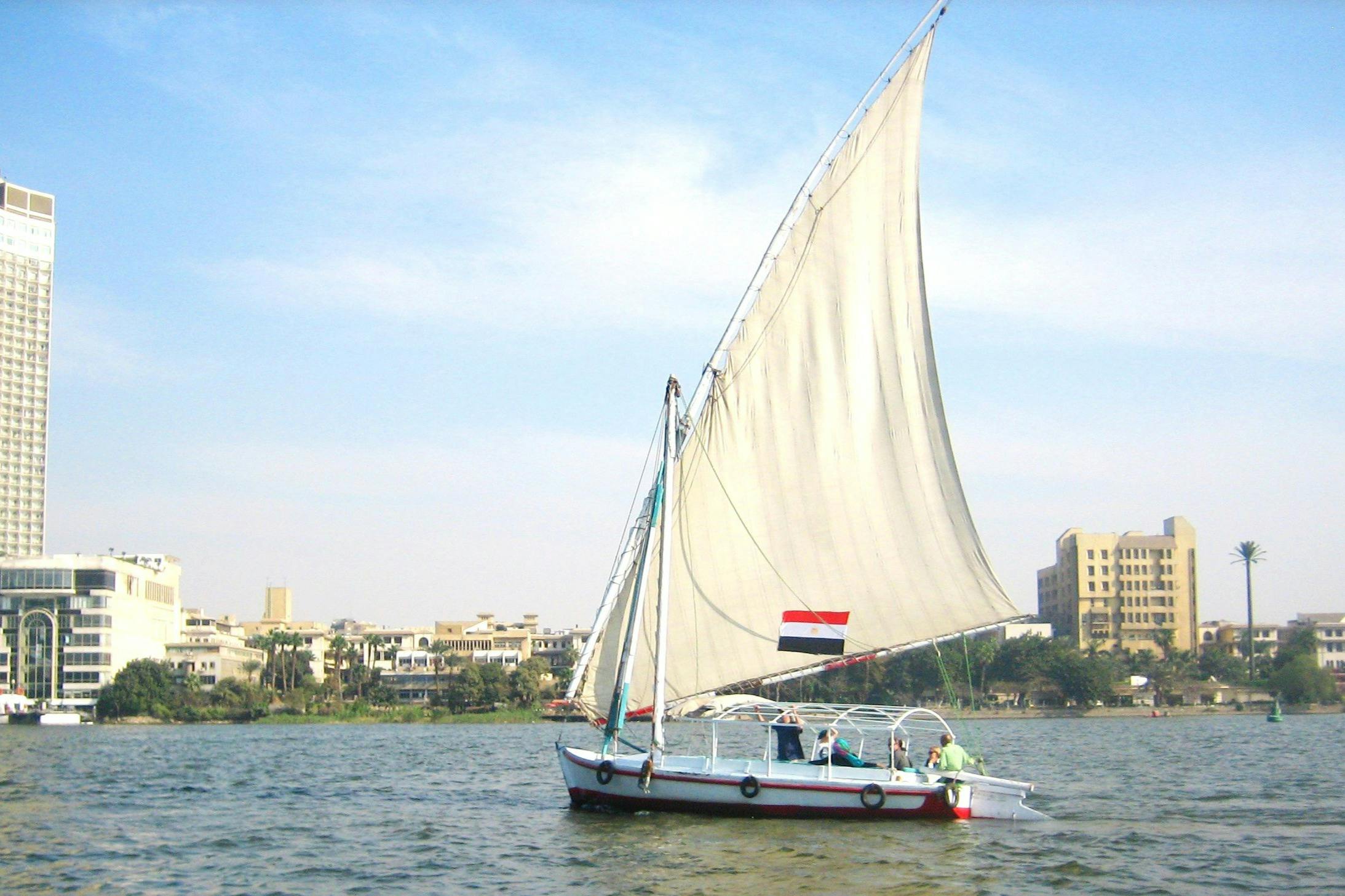 Ένα ιστιοφόρο με ένα μεγάλο λευκό πανί σε ένα ποτάμι, με μια σημαία, με κτίρια και δέντρα ορατά στο παρασκήνιο.