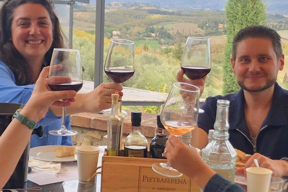 Four people sitting around a table outdoors, toasting with glasses of red wine. Scenic landscape visible in the background.
