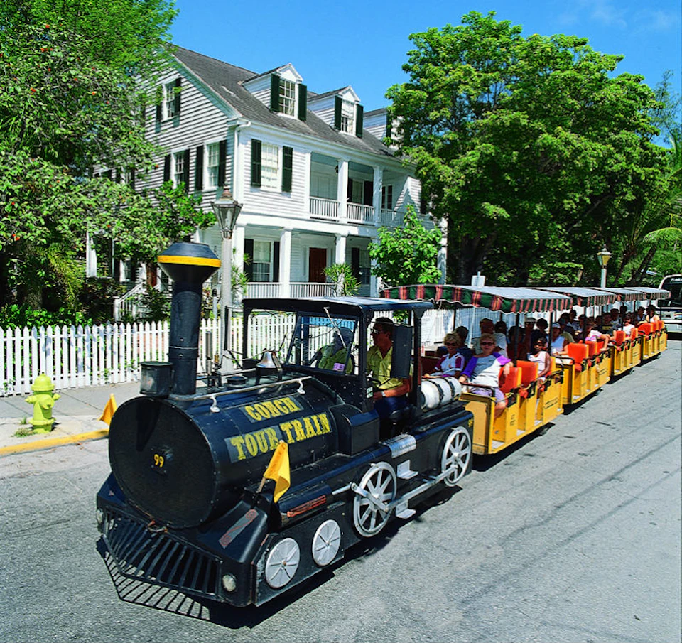 Tourists ride a small, black-and-yellow train labeled "Golden Tour Train" past a white house with green shutters and a picket fence.