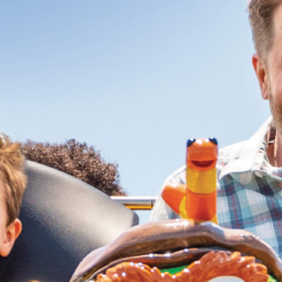 A man and boy excitedly ride a yellow rollercoaster themed with a green character and "I ♥ Trash" license plate.
