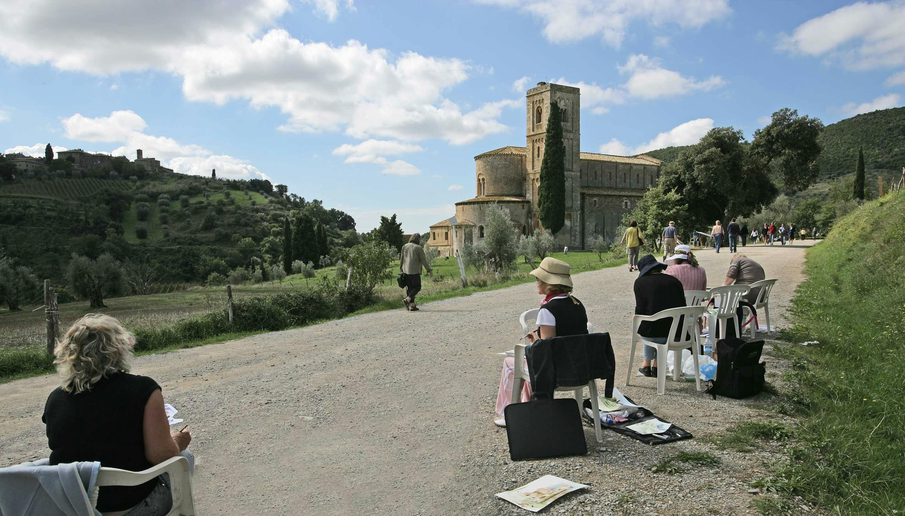 People sitting on chairs sketching in front of a historic church and enjoying a sunny day with scattered clouds.