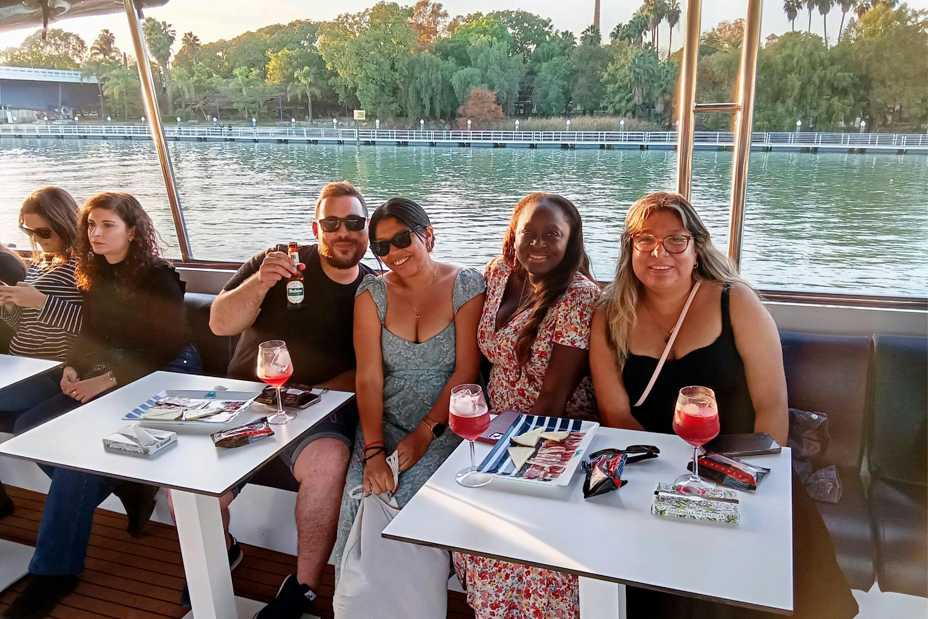 Een groep van vier mensen zittend aan een tafel met drankjes en hapjes op een boot, met water en bomen zichtbaar op de achtergrond.