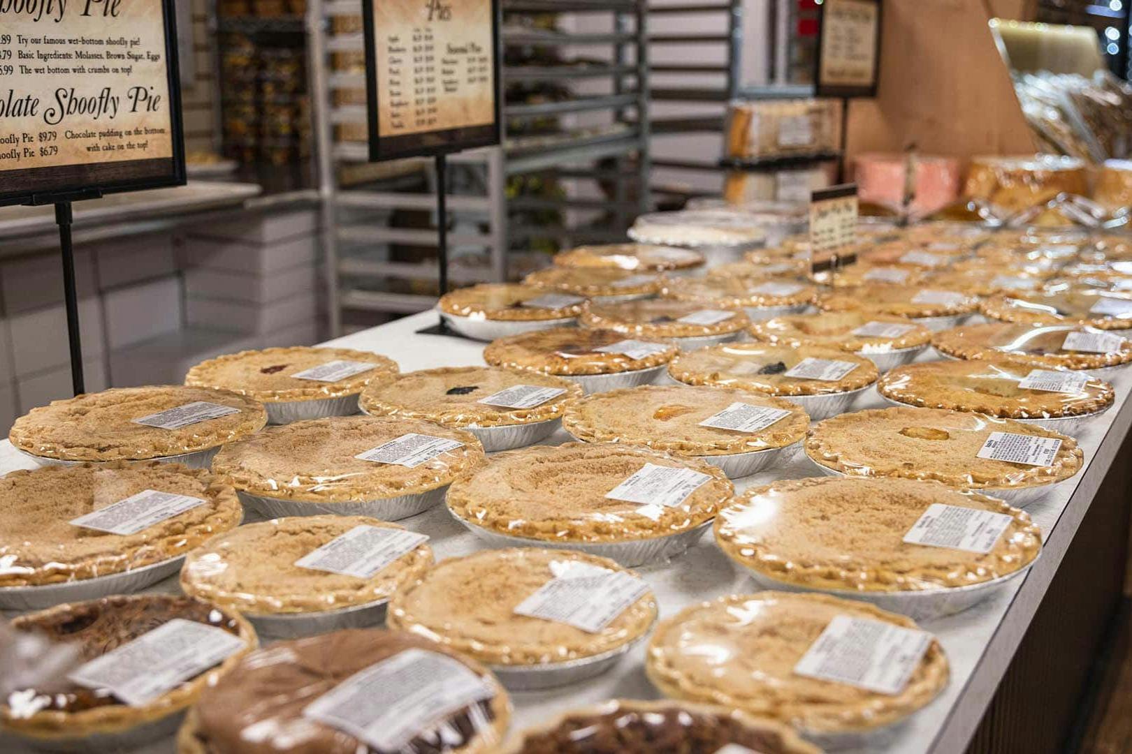 Rows of assorted pies in aluminum tins on a bakery store's display table with price signs in the background.