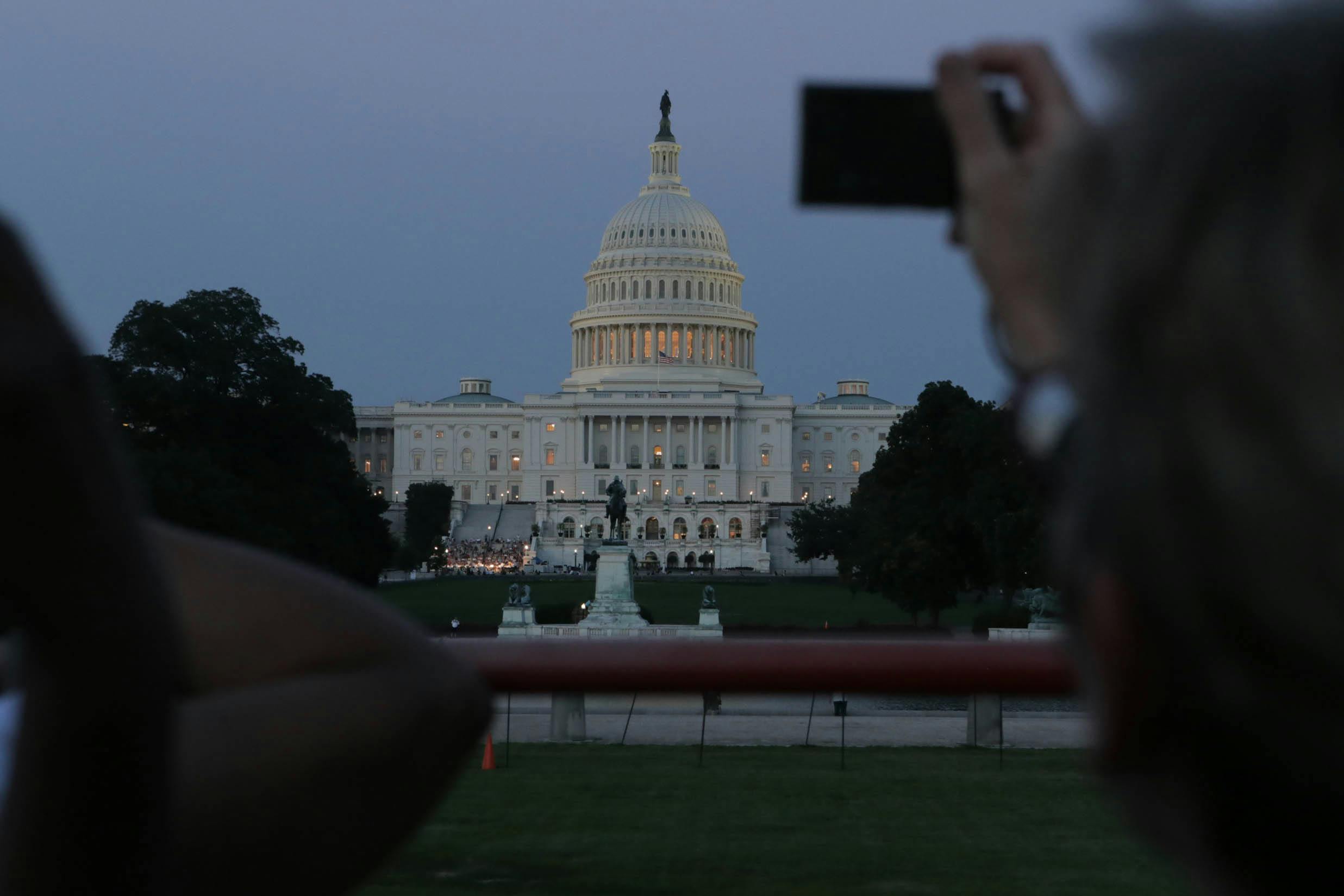 황혼이 질 무렵 미국 국회의사당 건물에 조명이 켜져 있으며, 전경에 앉아 있는 한 사람이 사진을 찍기 위해 휴대폰을 들고 있습니다.
