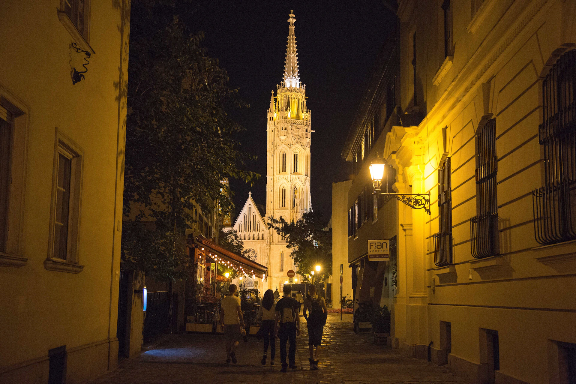 Group of people walking down a narrow, lamp-lit street towards a brightly illuminated church tower at night.