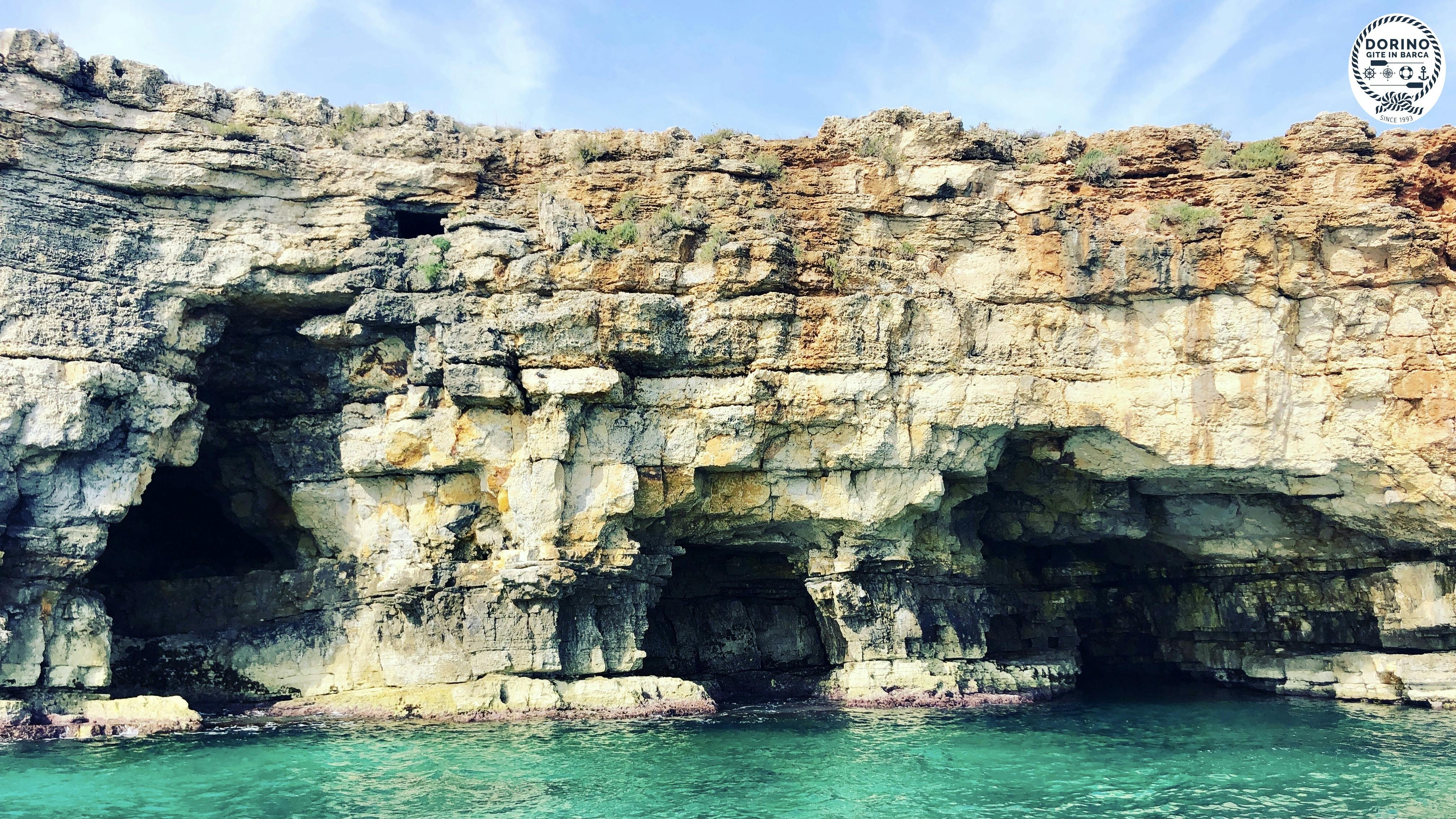 Scogliera rocciosa con grandi grotte a pelo d'acqua, delimitata da acque limpide e turchesi sotto un cielo parzialmente nuvoloso.