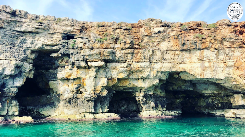 Rocky cliff with large caves at the water's edge, bordered by clear, turquoise water under a partly cloudy sky.