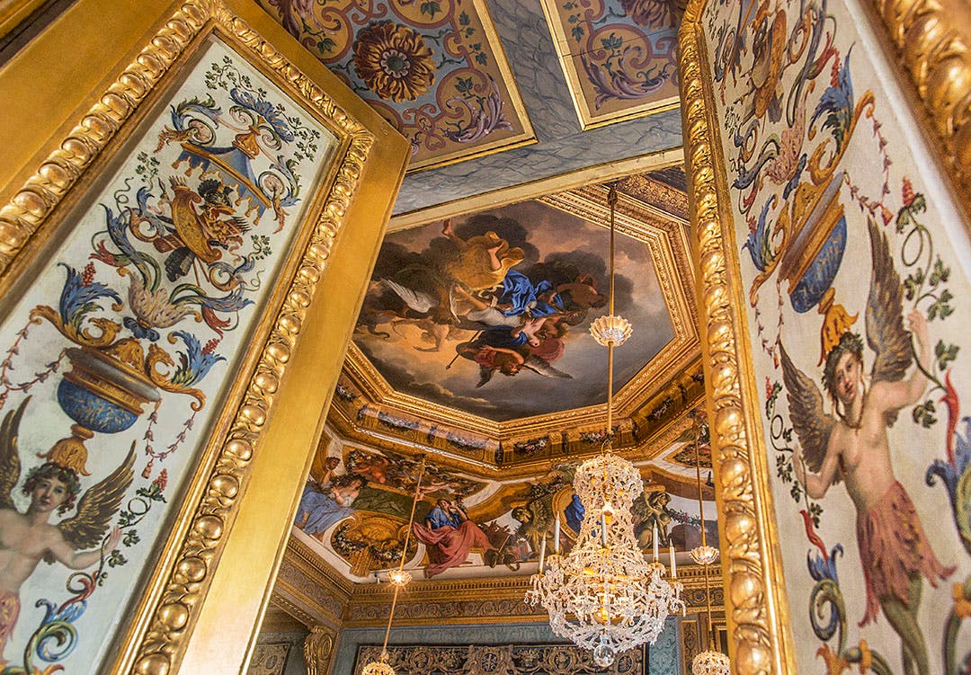 Ornate room with a painted ceiling depicting a mythological scene, golden-framed walls, and a crystal chandelier.