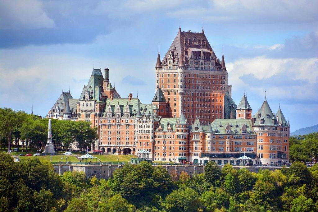 A large historic castle-like hotel with green roofs and a brick facade surrounded by trees under a partly cloudy sky.
