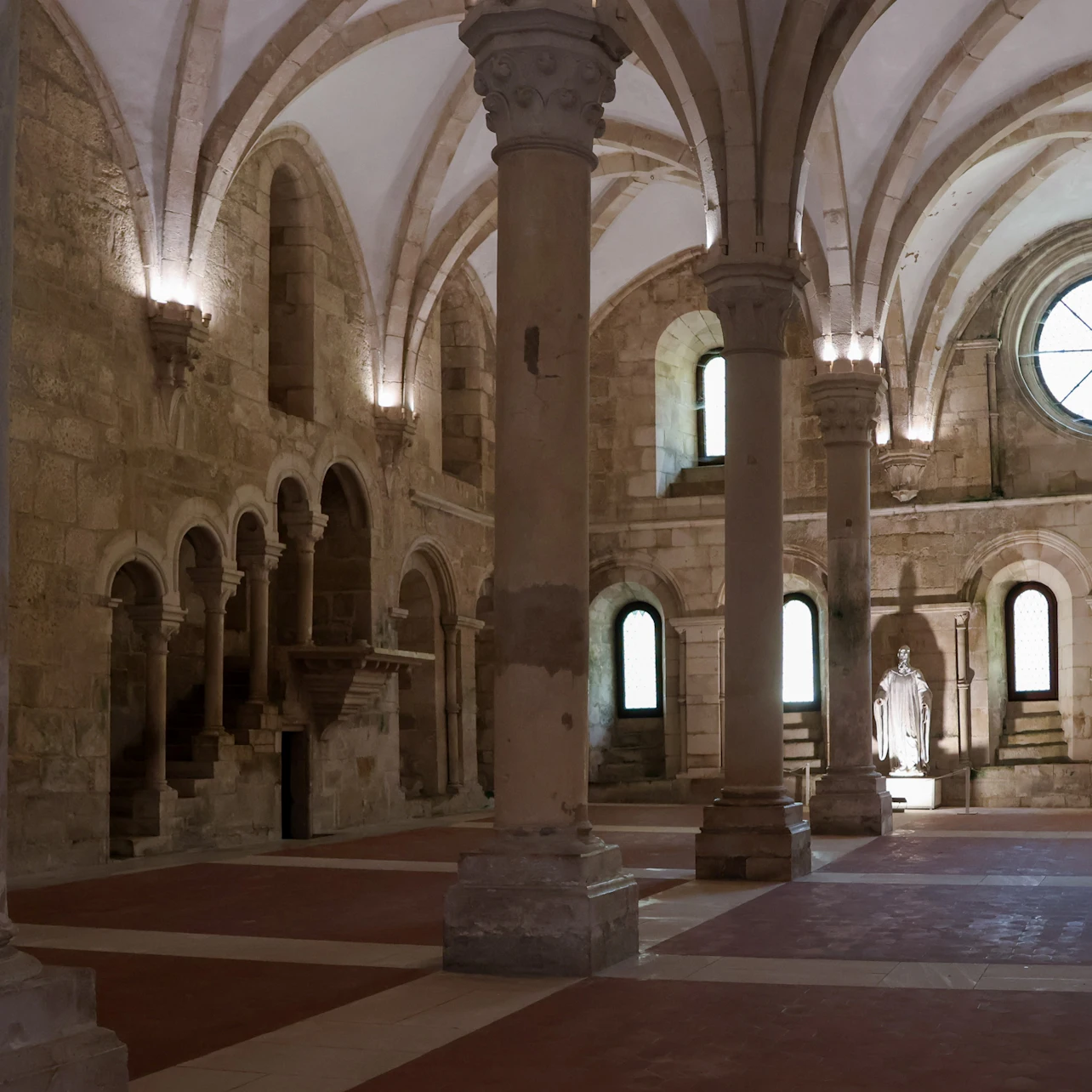 A historic stone hall with arches, columns, vaulted ceilings, and a statue in an alcove, illuminated by natural light.