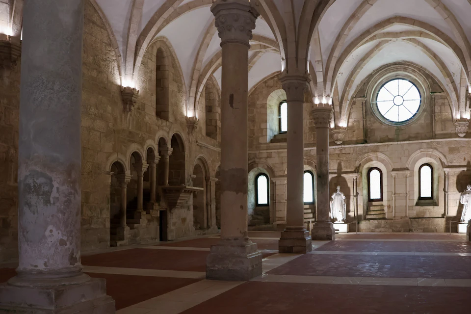 A historic stone hall with arches, columns, vaulted ceilings, and a statue in an alcove, illuminated by natural light.