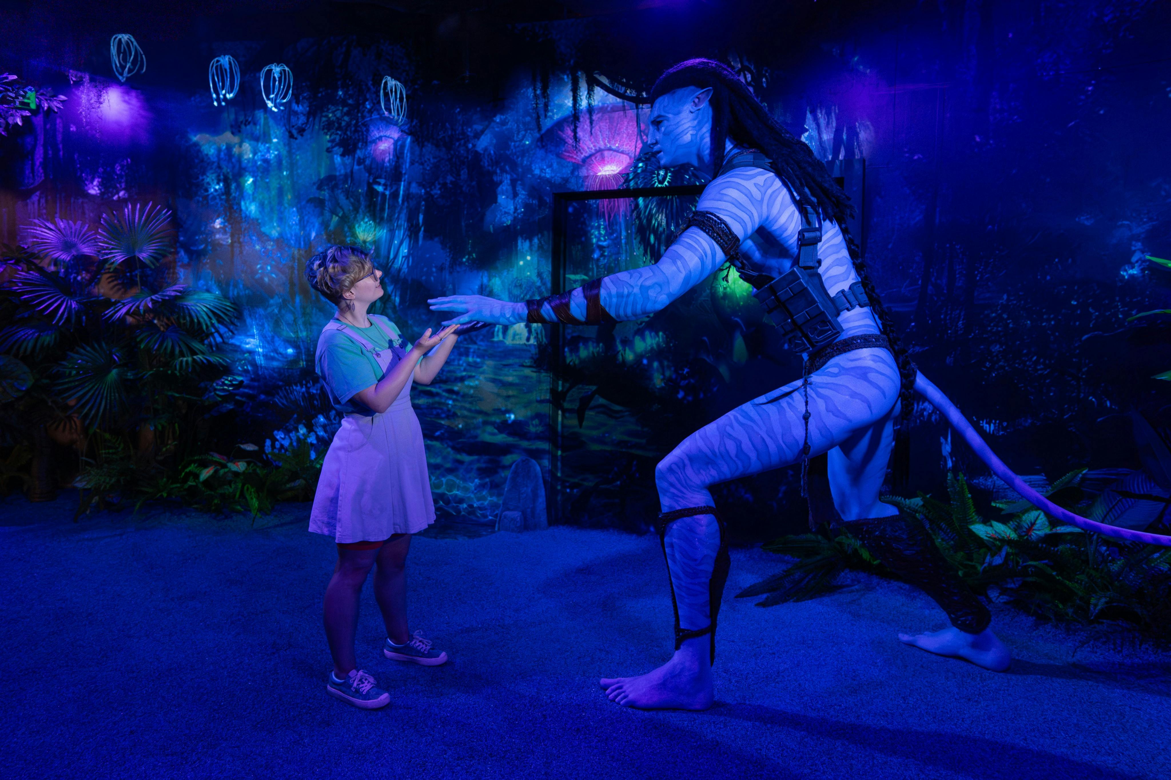 A woman interacts with a tall figure resembling a character from the movie Avatar in a themed, glowing environment.