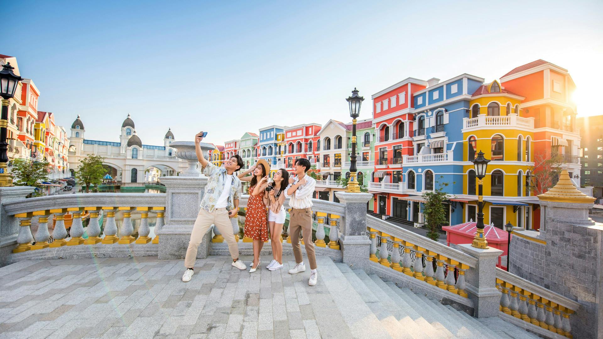Four people pose for a selfie on a decorative stone staircase in front of colorful buildings under a clear sky.