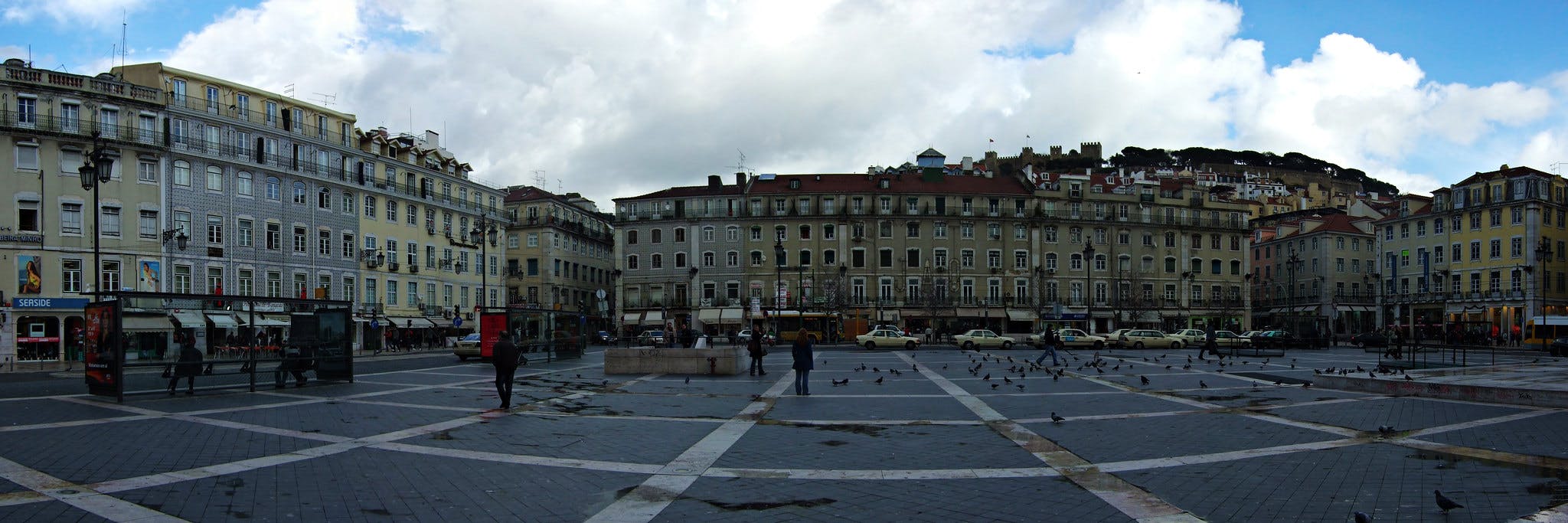 Ett stort öppet torg med duvor, omgivet av gamla flervåningsbyggnader med många fönster, butiker och några fotgängare.