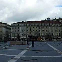 Ett stort öppet torg med duvor, omgivet av gamla flervåningsbyggnader med många fönster, butiker och några fotgängare.