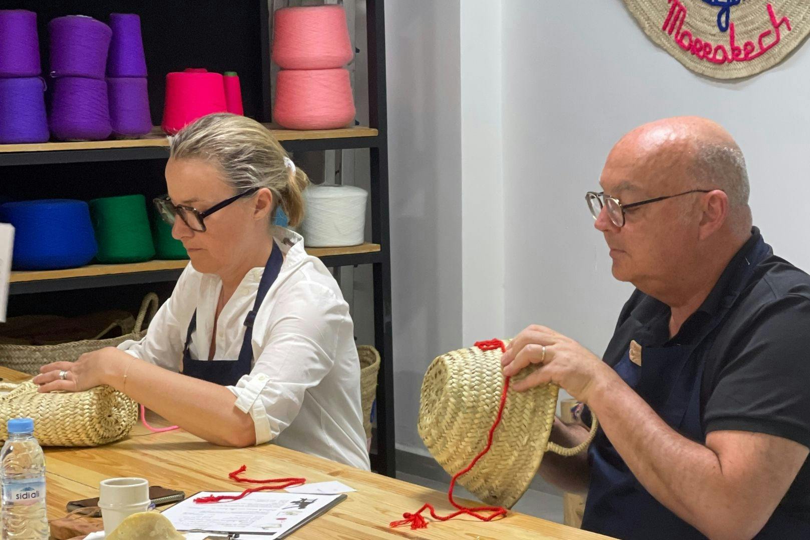 A woman wearing glasses and an apron works with straw baskets at a table, with colorful yarn on wooden shelves behind her.