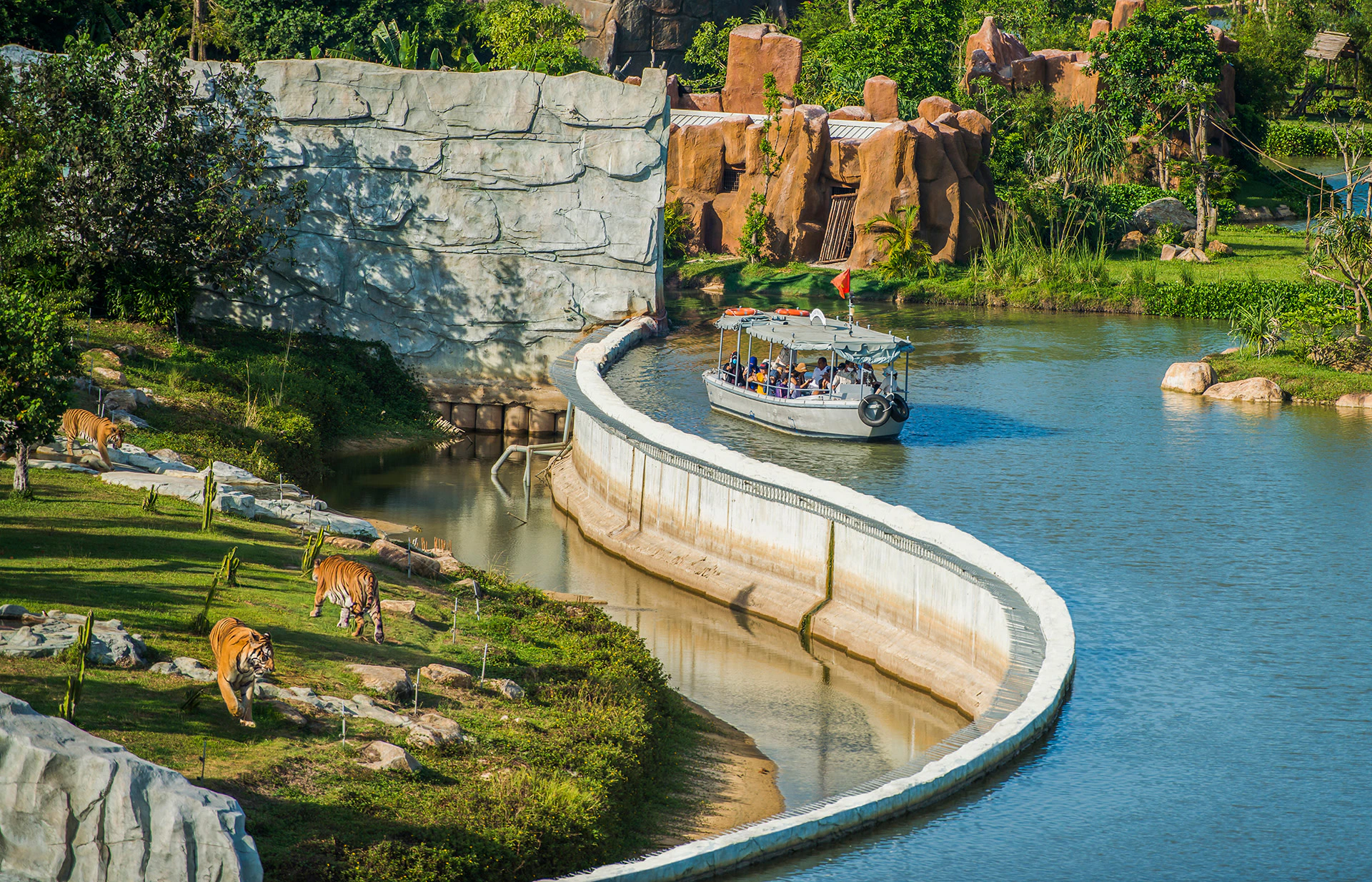 A boat with passengers sails along a winding waterway, with tigers and lush greenery visible on the bank nearby.
