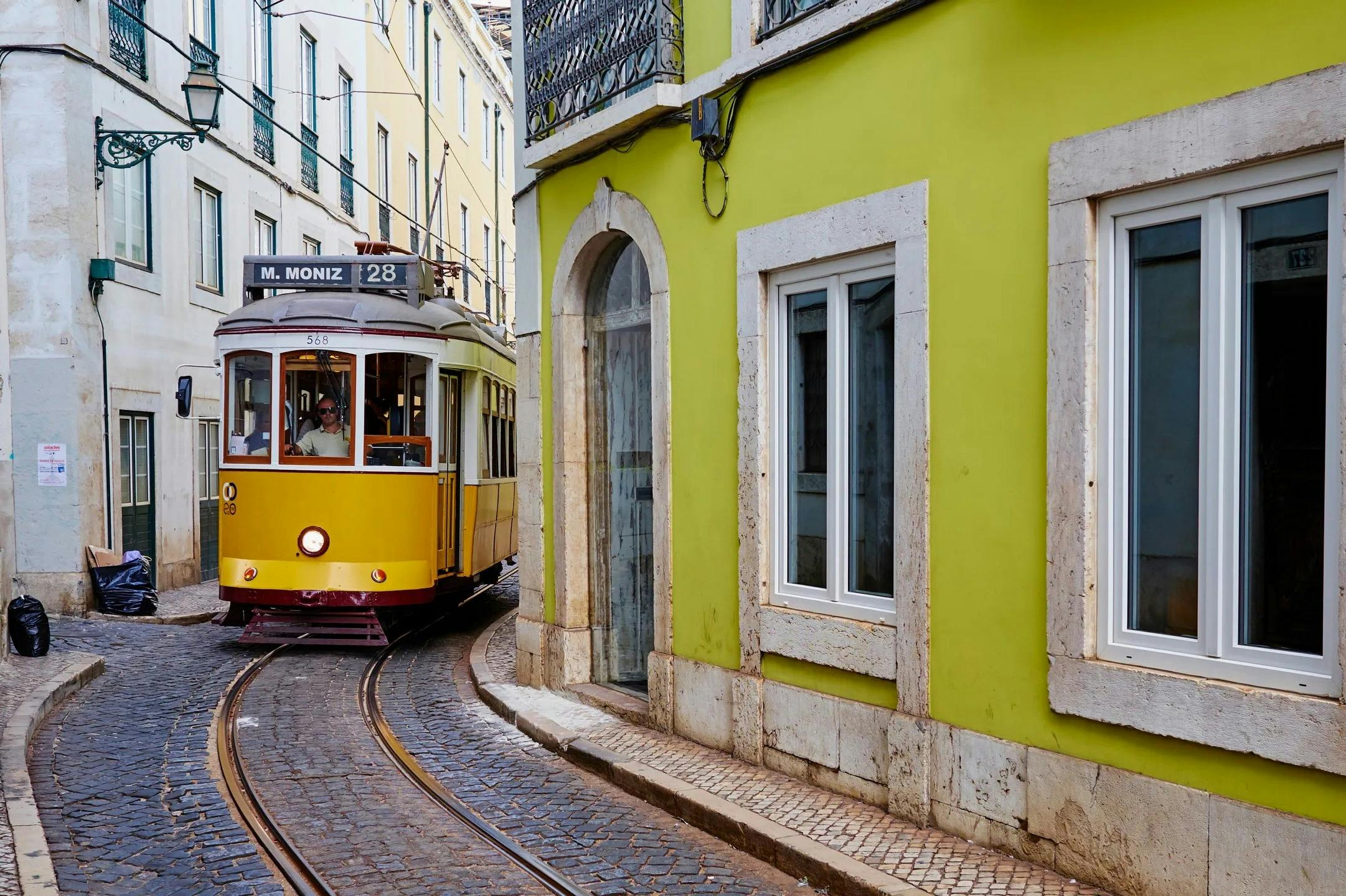 「28 M. Moniz」と記された黄色い路面電車は、白いトリミングの窓がある緑色の建物のそばの狭い石畳の通りにカーブしています。