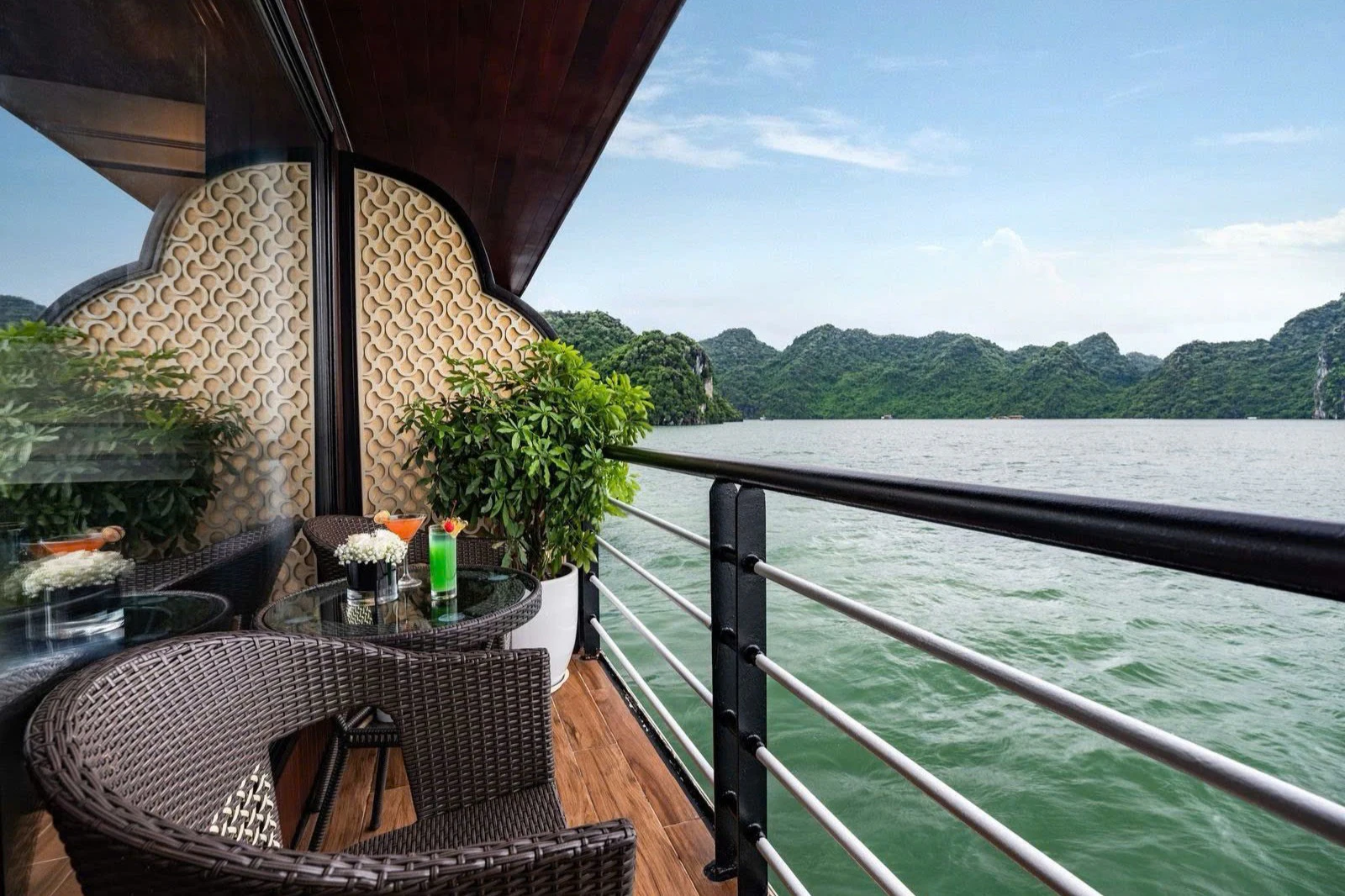 A boat balcony with wicker chairs, a table with drinks, potted plants, and a view of green hills across the water.