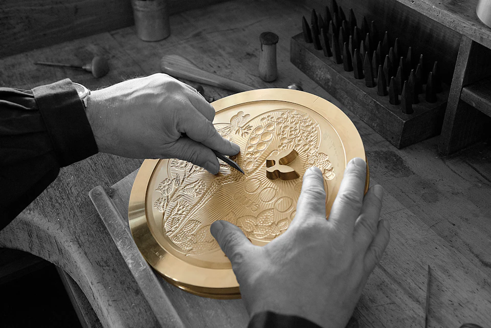 Hands engraving a detailed design onto a circular brass plate on a wooden workbench with several engraving tools nearby.