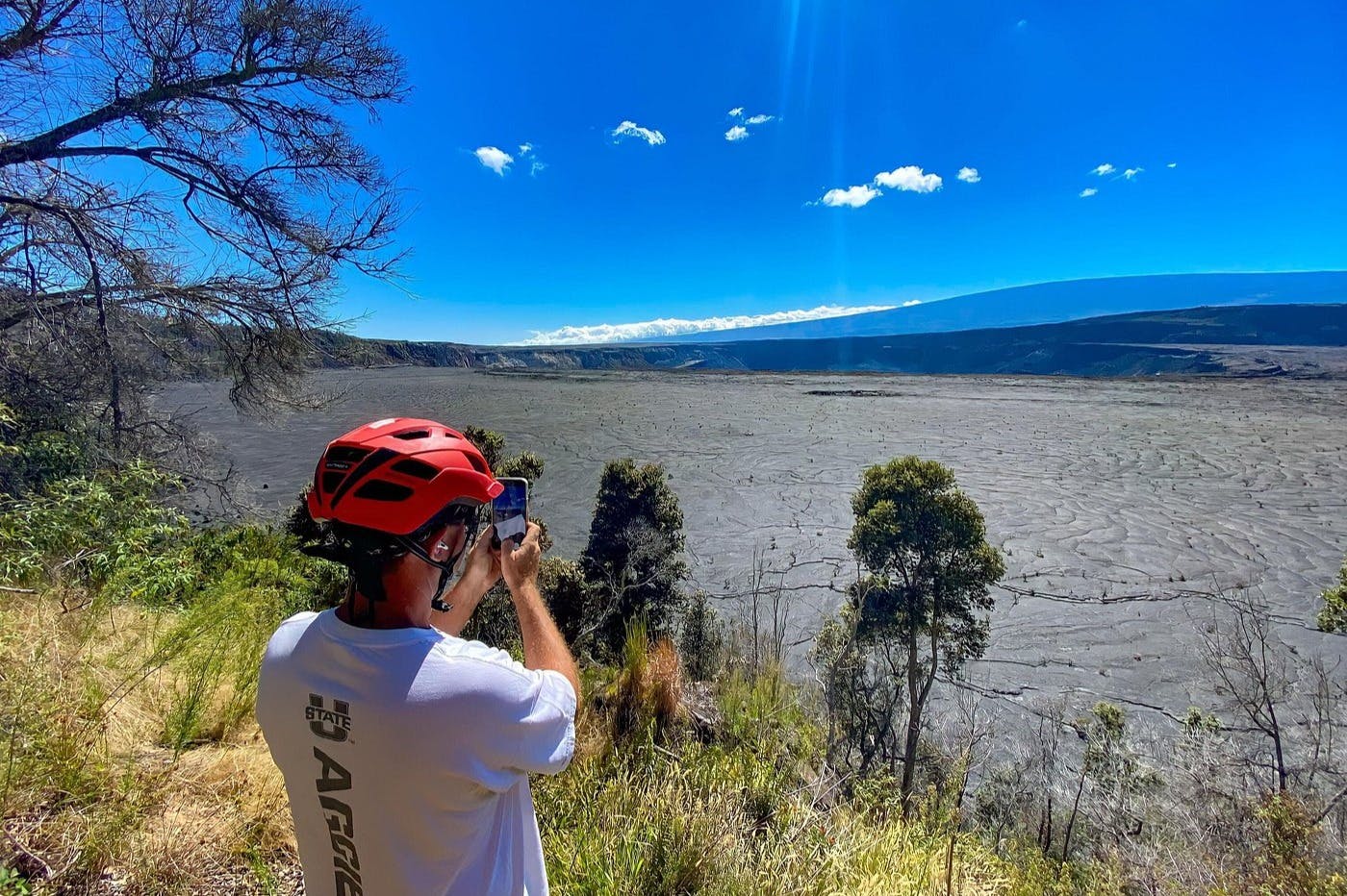 Osoba v červené helmě fotografující rozlehlou skalnatou krajinu s roztroušenými stromy a jasně modrou oblohou nad hlavou.