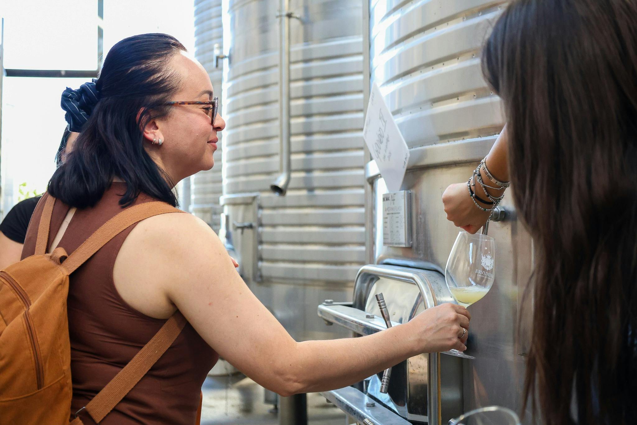 A woman with a brown backpack inspects a metal tank inside an industrial facility, with another person partially visible behind her.