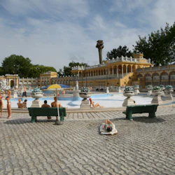 Människor kopplar av nära en torr fontän omgiven av gula byggnader och grönska, med bänkar och paraplyer runt omkring.