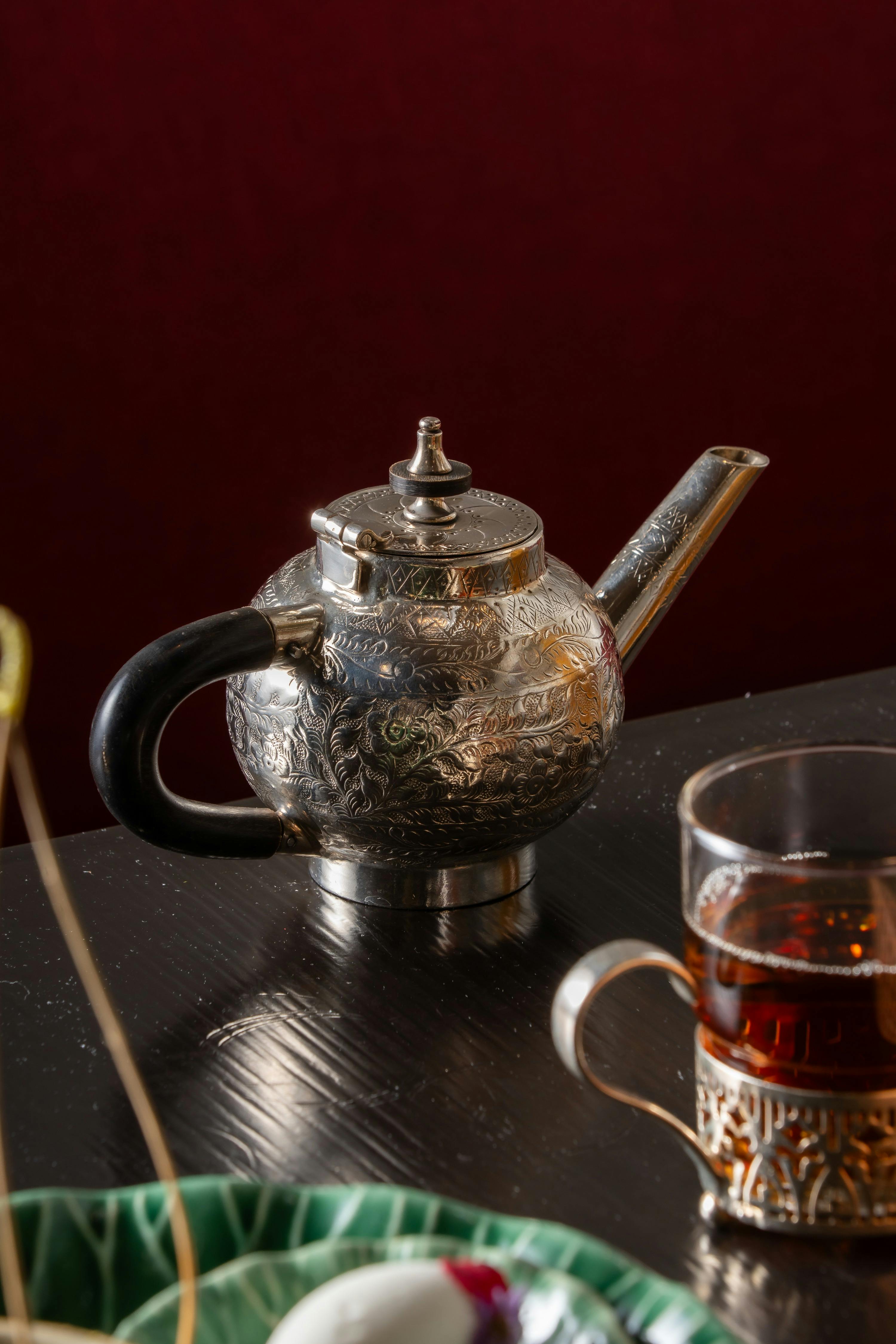A silver teapot with intricate patterns and a black handle sits on a dark table near a glass of tea with a metallic holder.