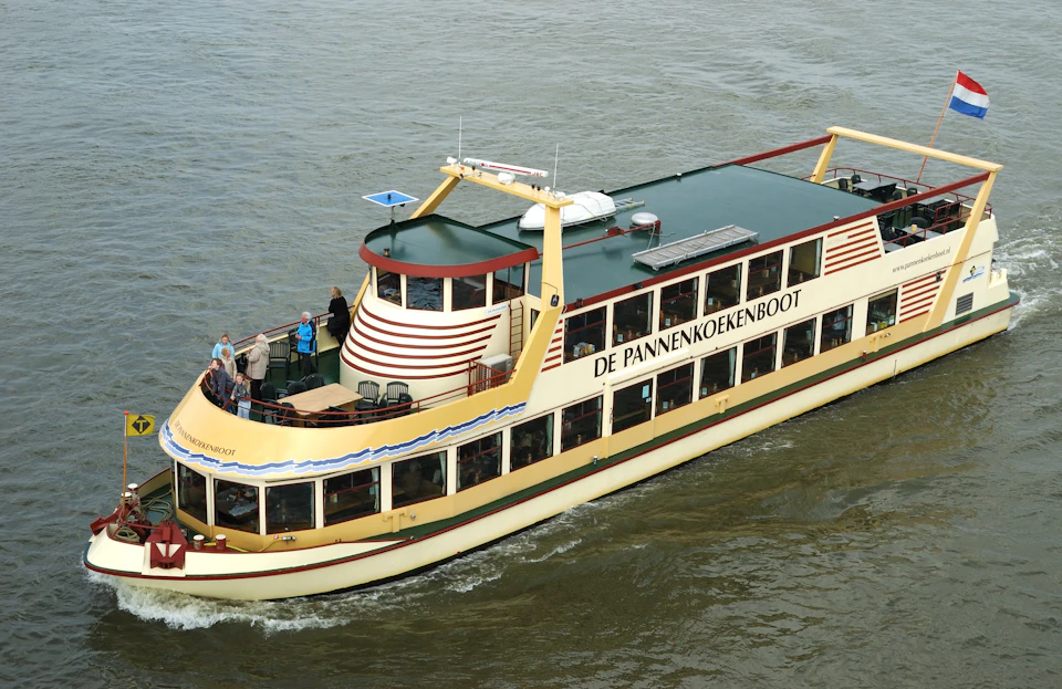 A boat named "De Pannenkoekenboot" cruises on water with passengers on its deck and in the windows.