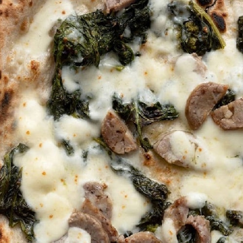 Close-up of a pizza with melted cheese, sausage slices, and leafy green vegetables on a thin, slightly charred crust.