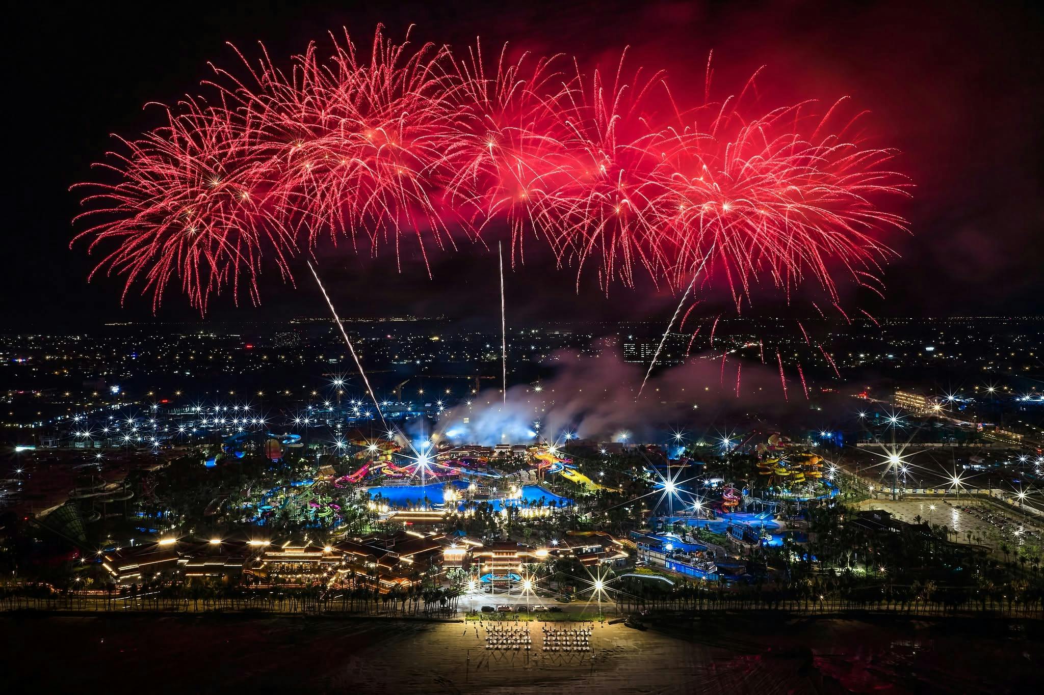 A nighttime aerial view of a brightly lit theme park with red fireworks exploding in the sky above.