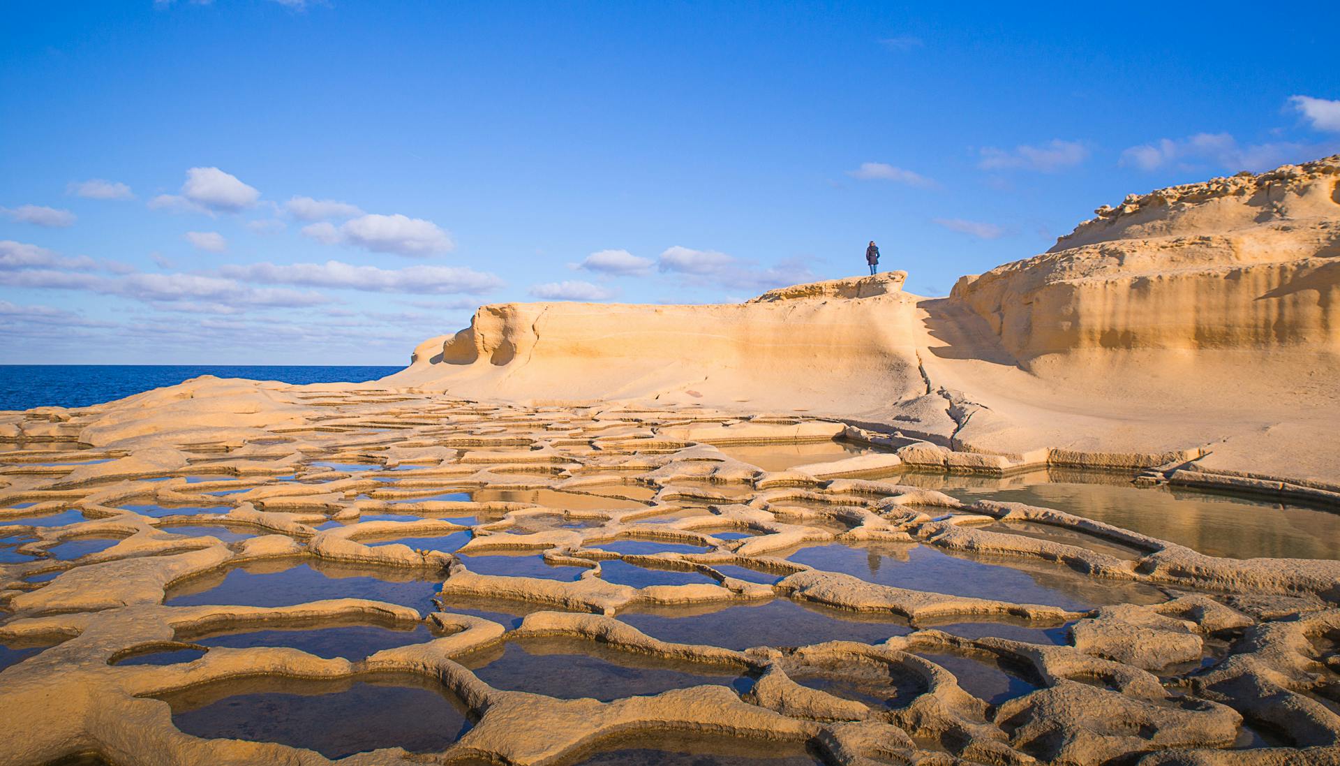 Una persona si trova su una scogliera di arenaria che sovrasta piscine di roccia erosa dal mare con uno sfondo di cielo azzurro e oceano.