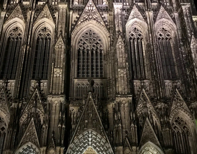 Gothic cathedral facade with intricate stone carvings, pointed arches, and large stained glass windows illuminated at night.