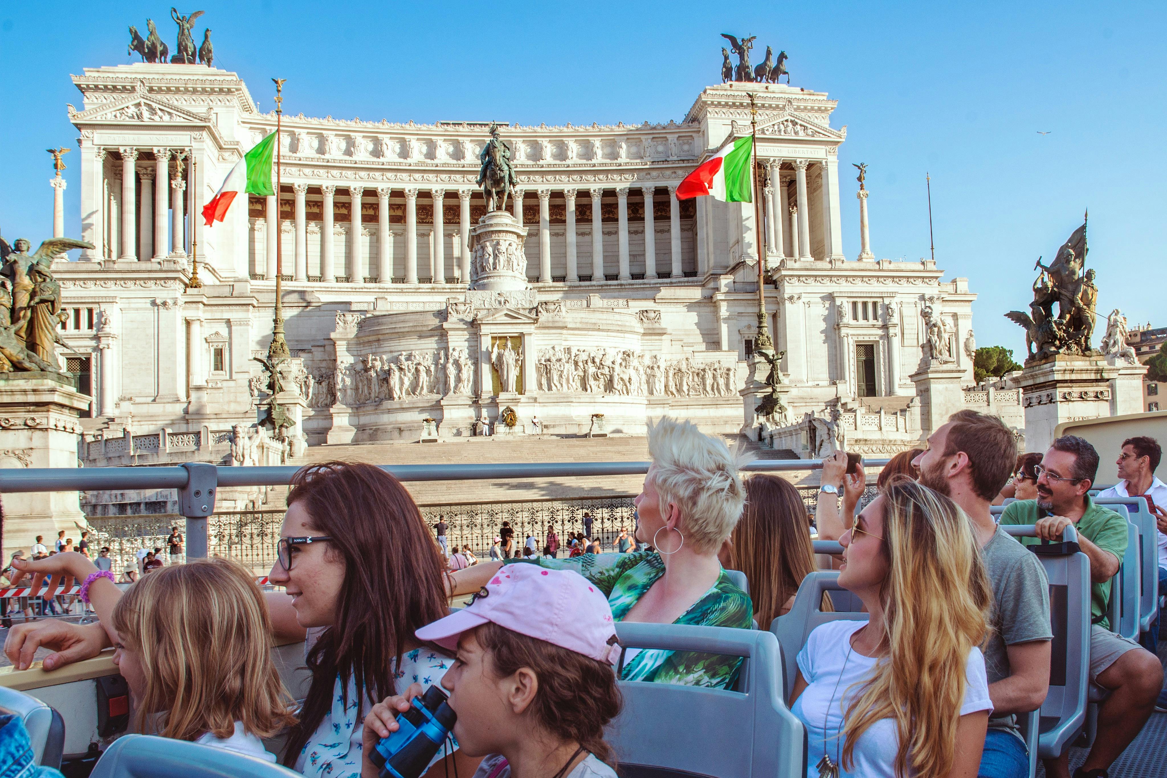 A group of people on a tour bus, looking at a grand historic building with an Italian flag, under a clear blue sky.