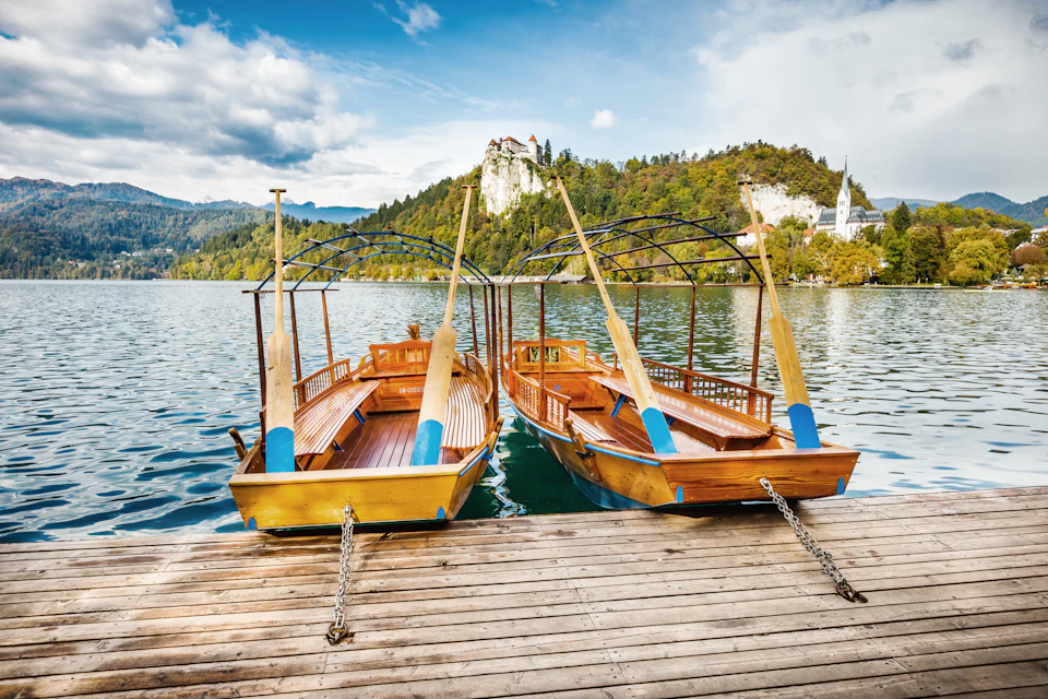 Two wooden boats with blue accents are moored at a dock on a lake, with a hill and buildings visible in the background.
