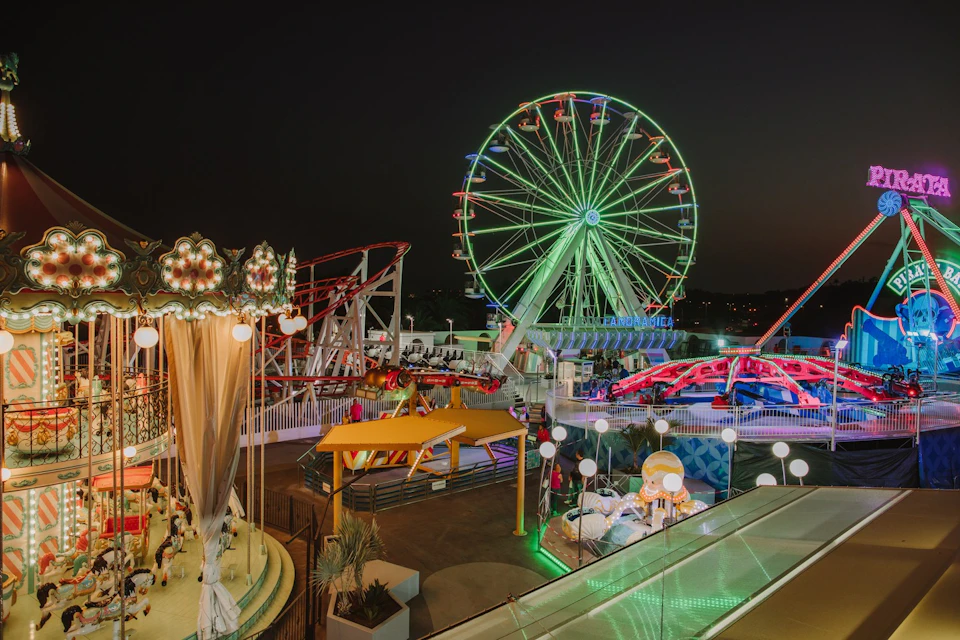 Amusement park at night with lit-up Ferris wheel, carousel, roller coasters, and colorful lights.