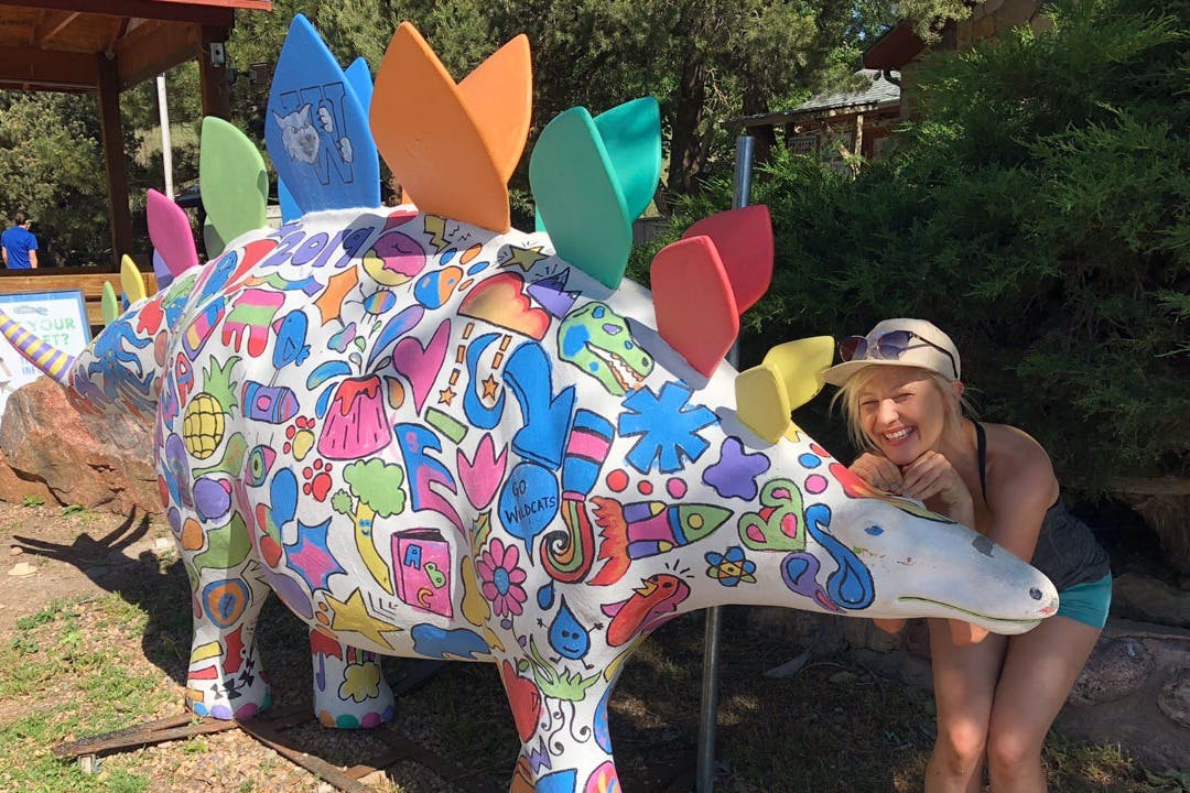 Una dona somrient al costat d'una estàtua d'estegosaure acolorida i decorada amb dissenys de dibuixos animats i plats vibrants. Arbres i arbustos al fons.