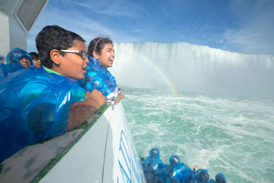 Children in blue ponchos smiling on a boat near a waterfall with a rainbow in the mist.