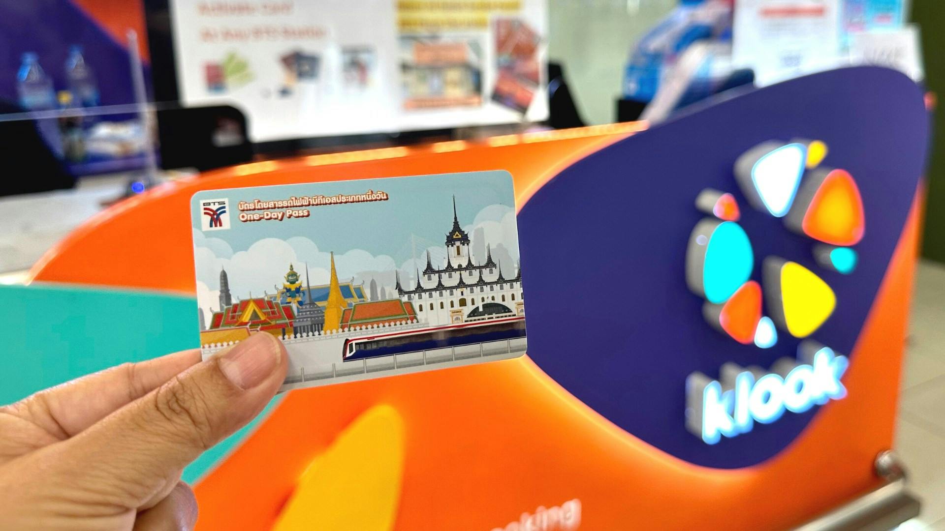 A hand holding a one-day pass card featuring a train and landmarks in front of a colorful, blurred background.