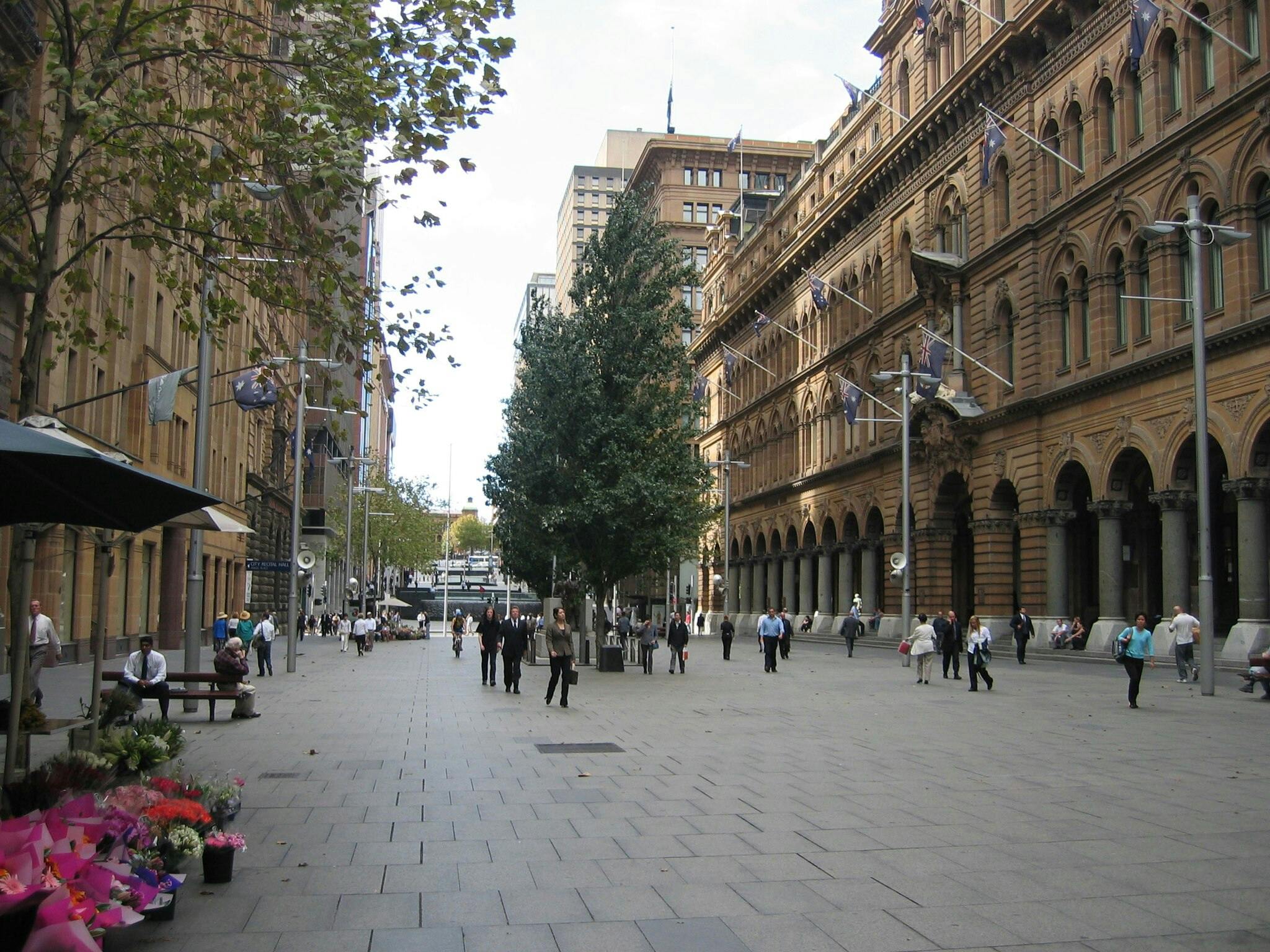 People walking along a wide, stone-paved street lined with historic buildings, trees, and a flower stall in the foreground.