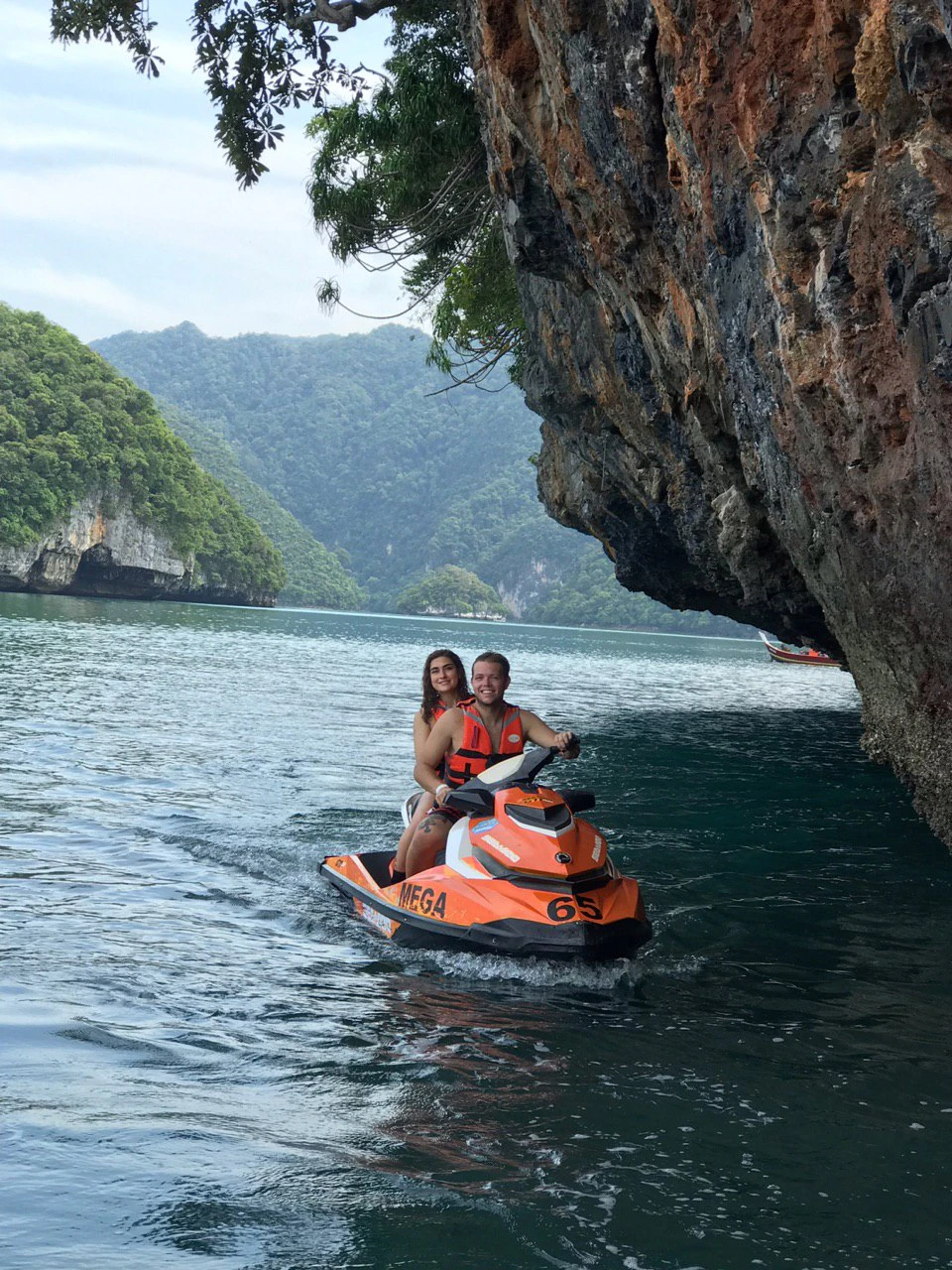 Two people riding a jet ski near a rocky cliff over calm water, with lush green hills in the background.