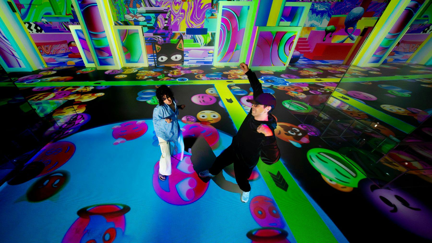 Zwei Personen interagieren in einem bunten Raum mit Projektionen von Emojis und abstrakten Grafiken an den Wänden und auf dem Boden.