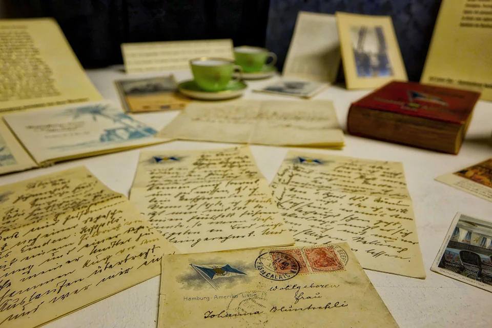 Vintage letters and postcards on a table, surrounded by green teacups, framed photos, and a red book.