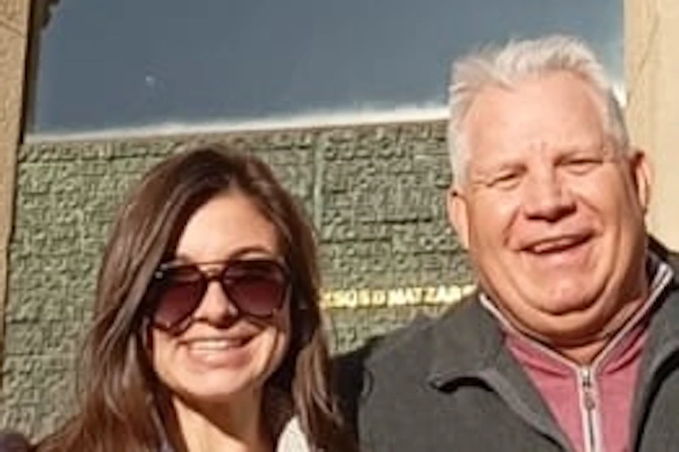 A group of four people smiling in front of a stone wall with sculpted figures. The scene is sunny and bright.