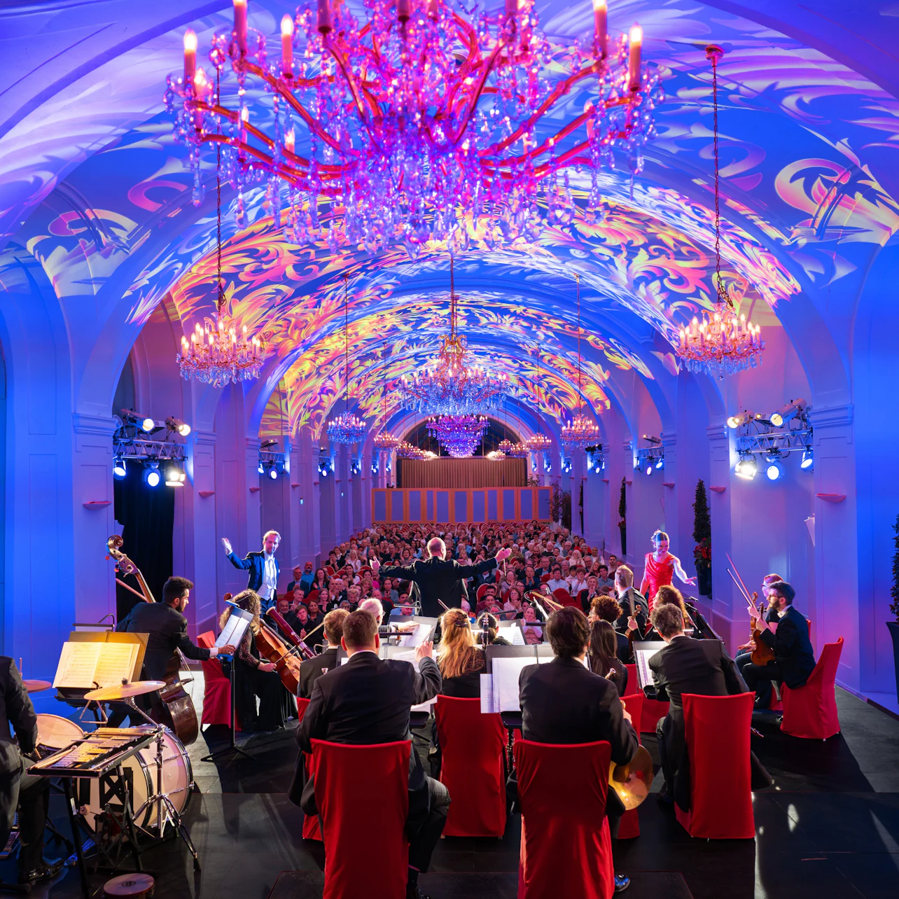 An orchestra performing in a grand hall with decorative lighting and chandeliers, facing an audience seated in rows.