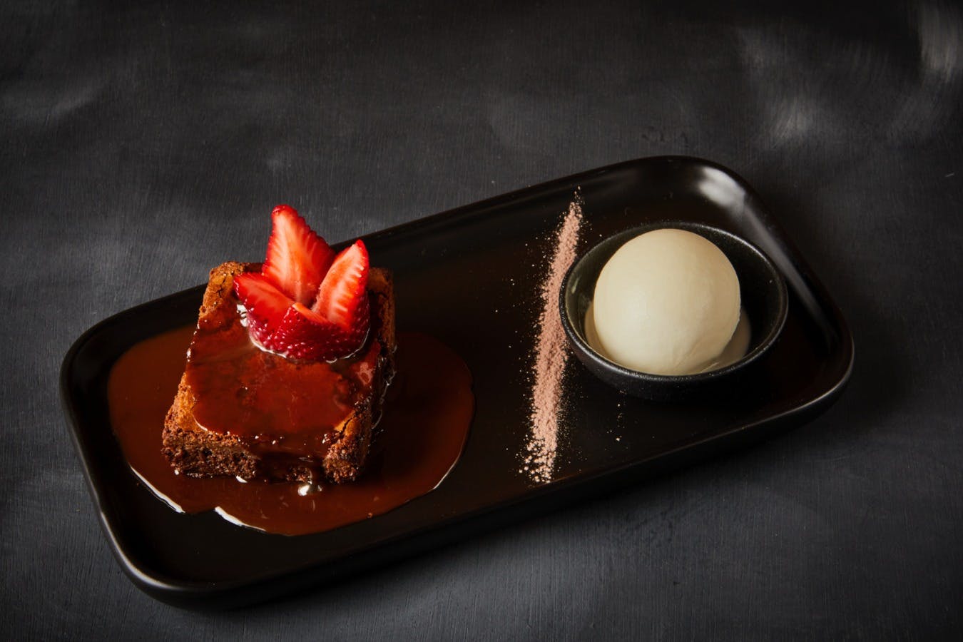 Ein Dessertteller mit einem Brownie, der mit Erdbeeren und Soße, einer Kugel Eis und Puderzucker bestreut ist.
